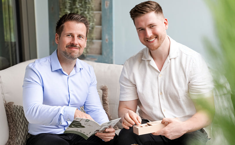Die Gründer von Naturtreu, Alex und Alex sitzen nebeneinander auf der Terrasse und lächeln in die Kamera. Sie halten den Produktkatalog und das Routineholz in ihren Händen.